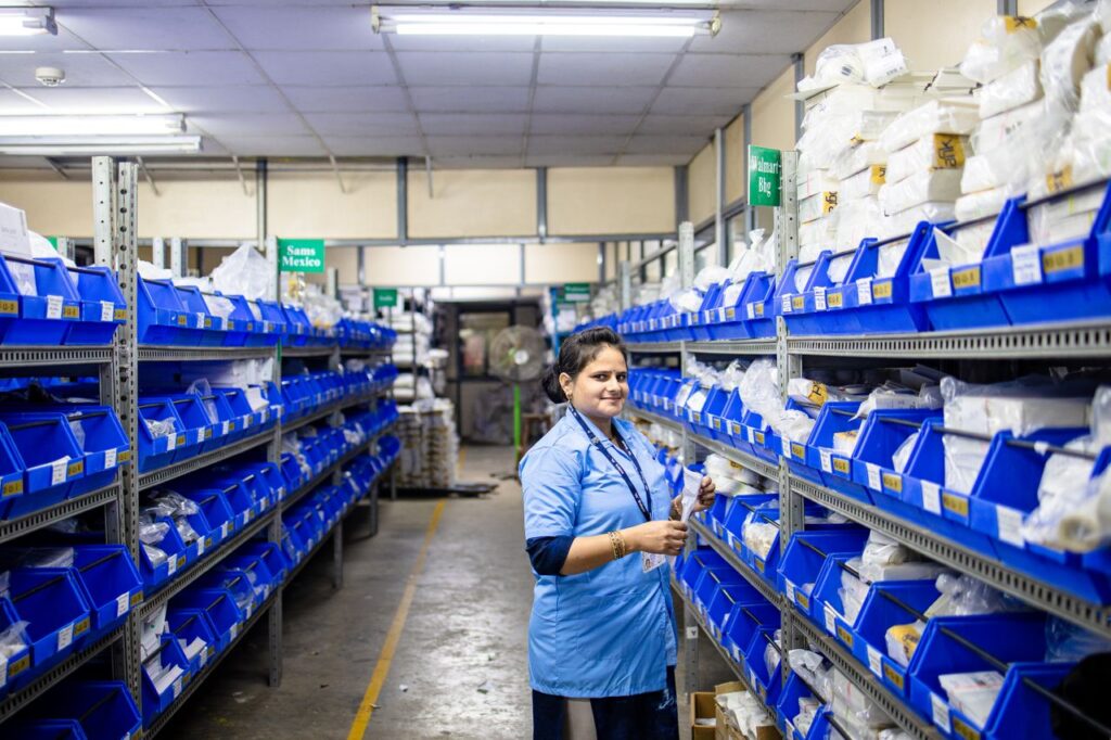 Female worker organizing inventory in a warehouse with blue bins and bright lighting.