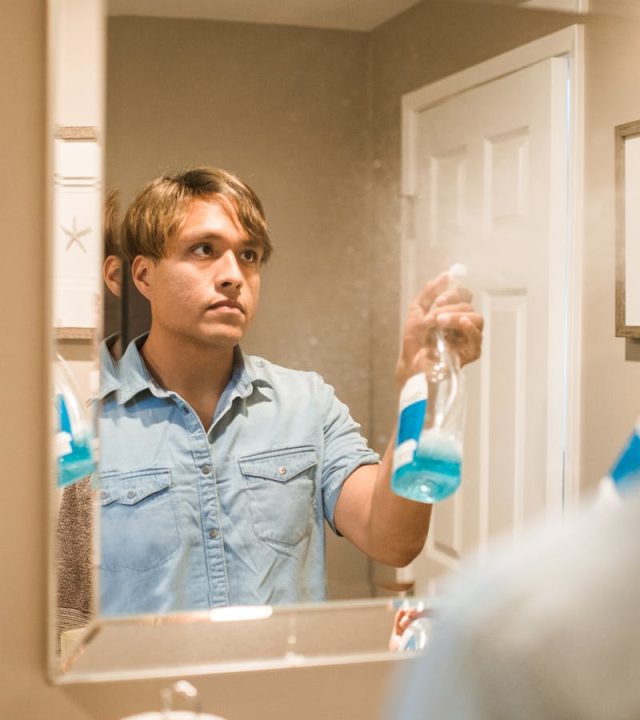 pexels-photo-5591457-5591457 Man cleaning bathroom mirror with blue spray bottle reflection in the frame.